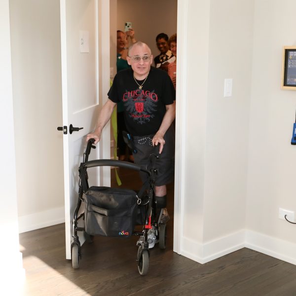 An elderly person with a walking aid enters a room, wearing a black T-shirt and shorts. A group of people is visible in the background, watching and smiling. The room has a picture frame and a white wall.