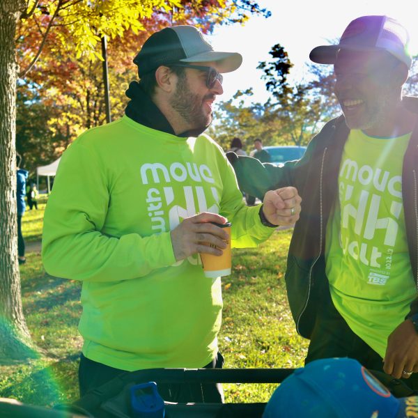 Two people in matching bright green shirts share a laugh in a park. One holds a coffee cup, and they are fist-bumping. Sunlight filters through autumn trees in the background, creating a cheerful atmosphere.