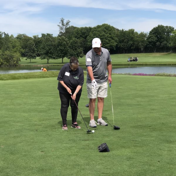 Two people on a golf course, with one person showing the other how to hold a golf club. They stand on a grassy area near a pond, with golf equipment lying on the ground. Trees and a golf cart are visible in the background.