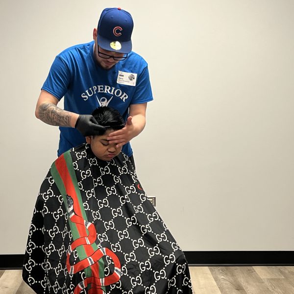 A person wearing a blue shirt and a cap is giving a haircut to another person who is seated and covered with a black Gucci cape. The barber is wearing black gloves and the setting appears to be a clean, modern barbershop with light-colored flooring.