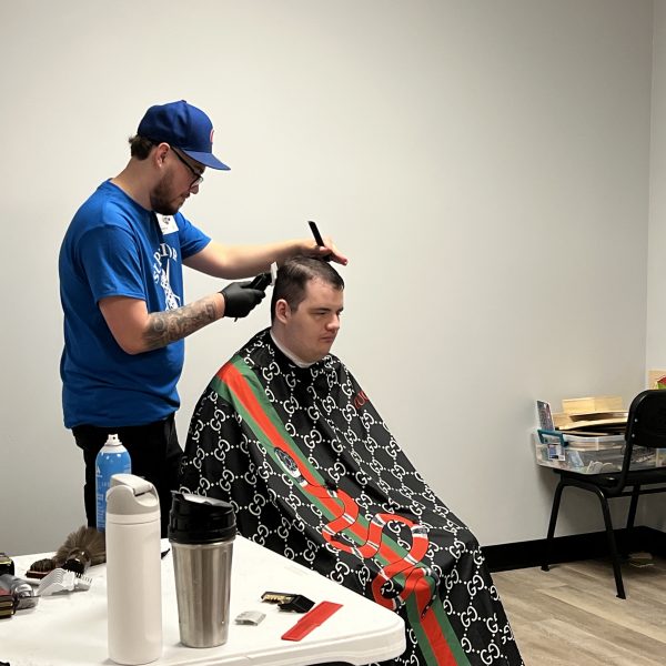 A barber in a blue shirt and cap is giving a haircut to a seated customer who is draped in a black, red, and green barber cape. A man in glasses and a green sweater stands nearby, watching. Various barber tools and products are on a table in the foreground.