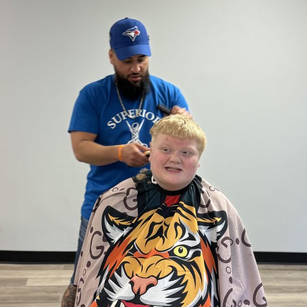 A barber wearing a blue cap and t-shirt is giving a young boy with light-colored hair a haircut. The boy is smiling and wearing a barber cape that features a large, colorful tiger design. The setting appears to be a modern barber shop with light wooden flooring.