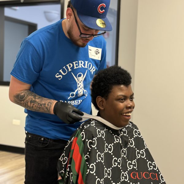 A barber wearing a blue "Superior Barber" t-shirt, blue cap, and gloves is trimming the hair of a smiling young boy who is covered with a Gucci barber cape. They are in a modern barbershop with a bright and clean interior.