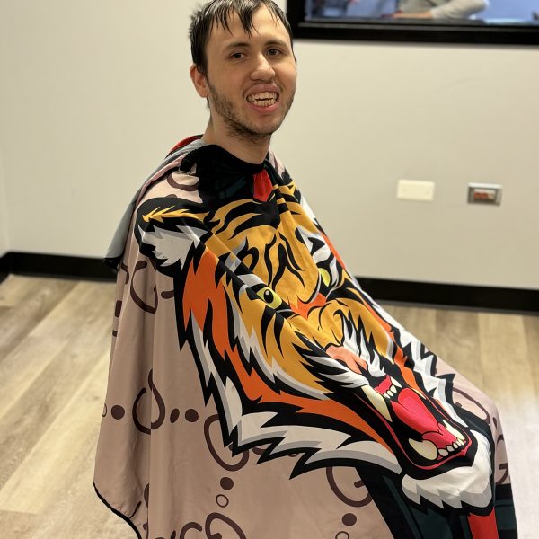 A man with wet hair smiles while sitting in a barber's chair, wearing a colorful cape featuring a tiger image. The room has light-colored walls and wooden flooring. A window in the background shows a child and some adults in another area.