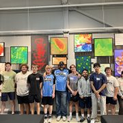 A group of people posing in front of a colorful art display. They are standing in a line and smiling. The artwork consists of abstract and vibrant paintings in various styles and colors. The setting appears to be an indoor gallery or exhibition.