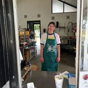 A person wearing a green apron and white t-shirt stands inside a food kiosk, smiling at the camera. The counter has a tip jar and a menu visible. The interior shows kitchen equipment and supplies.